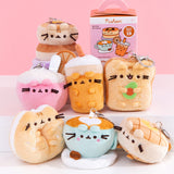 Front view of Pusheen Breakfast Surprise Plush assortment. Plush keychains sit on multi-level white pedestal in front of a pink background. Keychains feature food items including a pancake stack, cereal bowl, glass of orange juice, avocado toast, sesame bagel, coffee latte, and waffle. Accompanying the seven characters of breakfast Pusheens is the mystery box packaging for the Pusheen Breakfast Surprise Plush