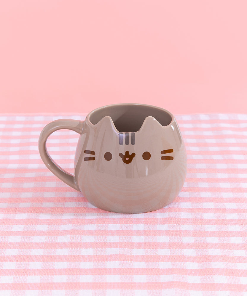 Pusheen Character Mug sits on a pink and white plaid surface. The grey mug has two ears to mimics Pusheen the Cat's face. On the front of the mug, is a printed face with brown whisker, eyes, and mouth details with grey stripes in between the ears.