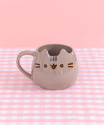 Pusheen Character Mug sits on a pink and white plaid surface. The grey mug has two ears to mimics Pusheen the Cat's face. On the front of the mug, is a printed face with brown whisker, eyes, and mouth details with grey stripes in between the ears.