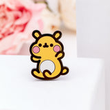 The Cheek pin sitting on a white pedestal, another white pedestal and a few pink peonies in the background. The pin features Cheek the hamster sitting, feet and hands raised upwards.