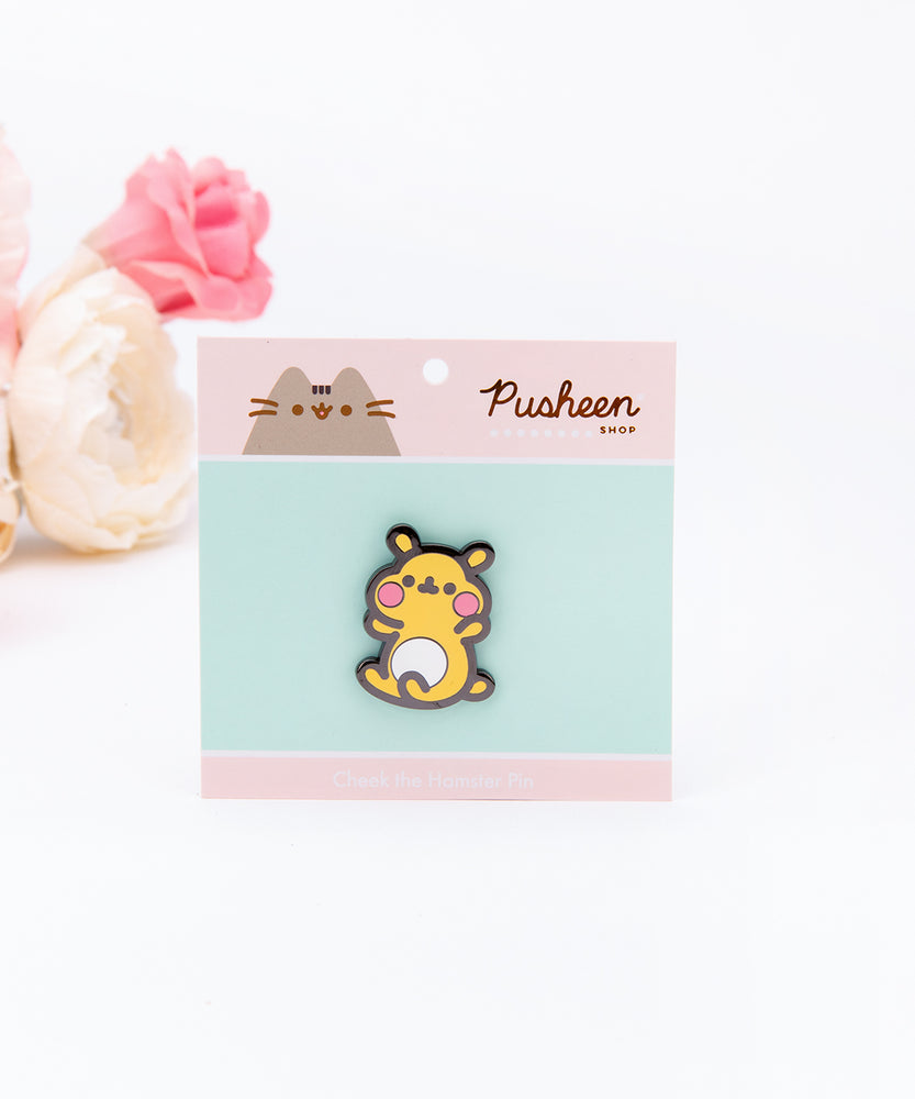The Cheek pin in it’s packaging, laying on a white floor, a pair of pink and white peonies partially visible in the top left corner. The packaging is a square cardboard backing, a mint square in the middle and pink banners on the top and bottom. The top banner features the Pusheen Shop logo, and the bottom banner says ‘Cheek the Hamster Pin’ in white.