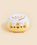A round Pusheen plush that resembles a cinnamon roll bun with frosting on top with a cinnamon swirl. Pusheen's ears are covered by the frosting, and the three light brown embroidered stripes in between her ears peek out from the frosting.