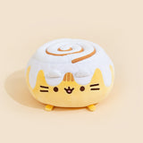 A round Pusheen plush that resembles a cinnamon roll bun with frosting on top with a cinnamon swirl. Pusheen's ears are covered by the frosting, and the three light brown embroidered stripes in between her ears peek out from the frosting.
