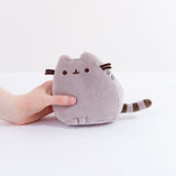 A model’s hand holding the Grey Mini Squisheen Plush in front of a white background. This plush is in Pusheen’s default colors.