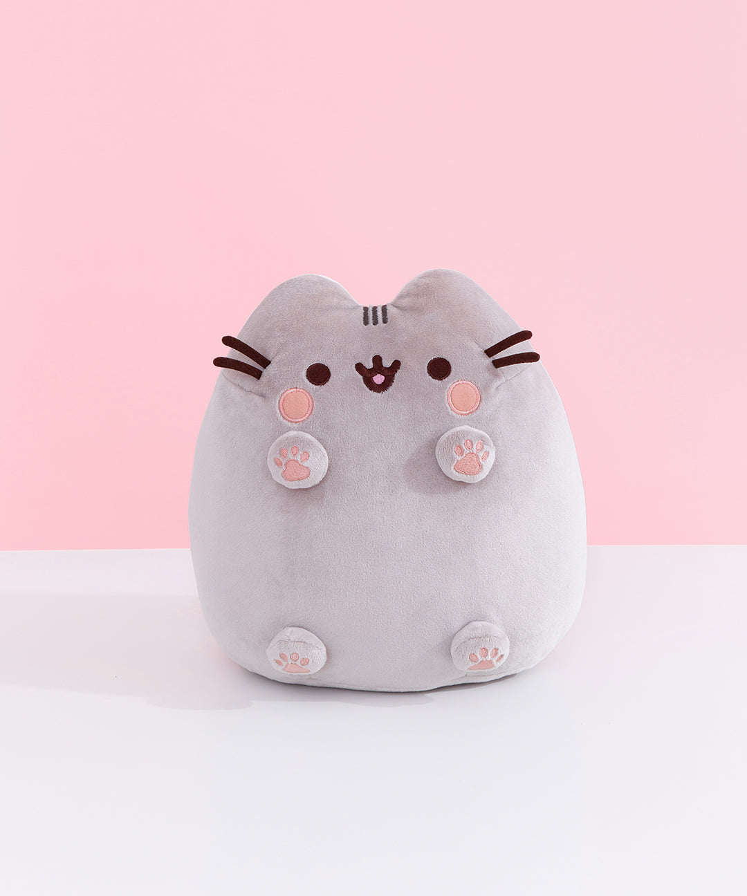 Front view of Classic Toe Beans Pusheen Plush. A blushing Pusheen the Cat shows off her pink toe beans and feet pads on her four paws that extend off her body. Her grey body is accentuated with her classic brown eyes, mouth, and whiskers.  