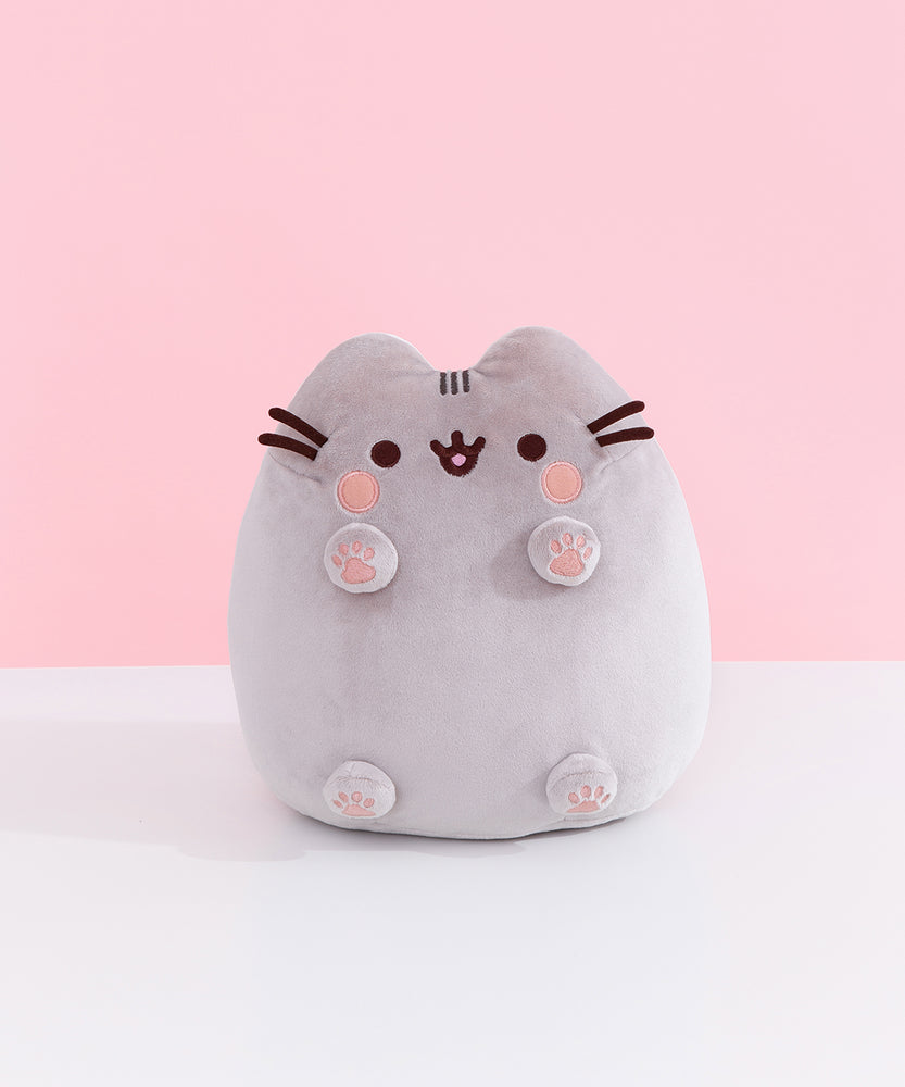 Front view of Classic Toe Beans Pusheen Plush. A blushing Pusheen the Cat shows off her pink toe beans and feet pads on her four paws that extend off her body. Her grey body is accentuated with her classic brown eyes, mouth, and whiskers.  