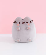 Front view of Classic Toe Beans Pusheen Plush. A blushing Pusheen the Cat shows off her pink toe beans and feet pads on her four paws that extend off her body. Her grey body is accentuated with her classic brown eyes, mouth, and whiskers.  