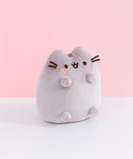 Right quarter view of Pusheen Toe Beans Plush. In this view, Pusheen’s whiskers and feet are shown extended from the plush body.  The plush sits atop a white table in front of a light pink backdrop.