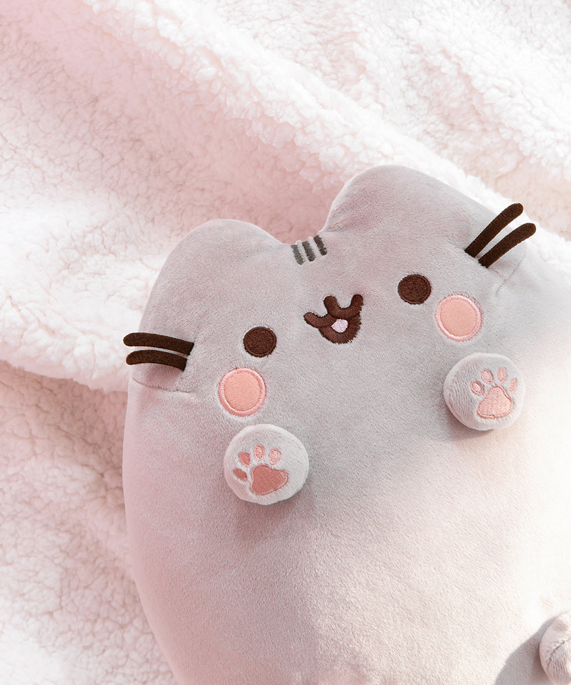   Close-up view of Classic Toe Beans Plush. Pusheen plush toy lays on a white fluffy surface. The front paws from the plush body while the pink toe beans are embroidered in a light pink thread. Pusheen’s light pink embroidered blush sits under her brown eyes. 