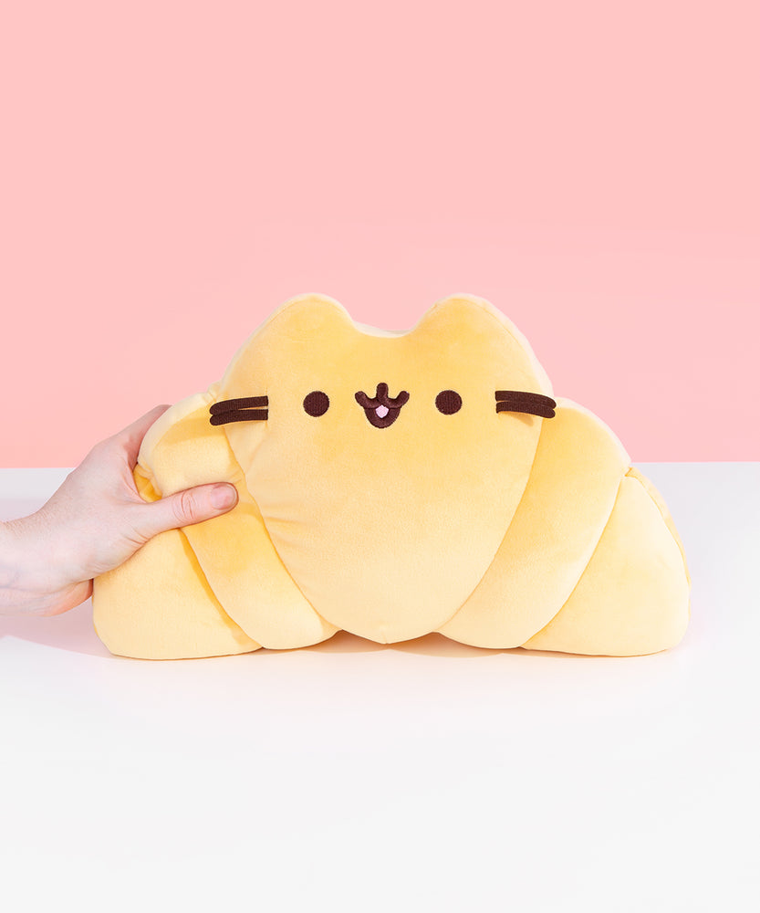 Model squeezes left side of Pusheen as a croissant-shaped plush. Plush has brown embroidery of Pusheen's eyes and mouth while brown whiskers extend off the croissant shape.