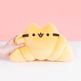 Model squeezes left side of Pusheen as a croissant-shaped plush. Plush has brown embroidery of Pusheen's eyes and mouth while brown whiskers extend off the croissant shape.