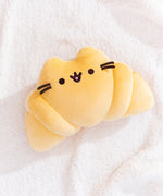 Croissant-shaped plush lays on a fluffy white surface. Pusheen's classic ears, whiskers, eyes, and mouth act as the face of the croissant-inspired plush.