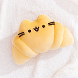 Croissant-shaped plush lays on a fluffy white surface. Pusheen's classic ears, whiskers, eyes, and mouth act as the face of the croissant-inspired plush.