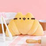 Front view of Croissant Pusheen Squisheen. Yellow plush features rolled sections towards the end of the plush to emulate croissant shape. Plush is surrounded by a brown and white bread basket, white tongs, a cream and brown rolling pin, and a brown coffee mug.