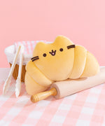 Croissant Pusheen Squisheen sits atop a beige rolling pin. The croissant-inspired plush toy is surround by a bread basket, tongs, a pink and white dish towel. The yellow plush sits on a pink and white plaid table cloth.
