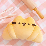 Croissant Squisheen lays flat on a white and pink checkered table cloth. The plush is surrounded by white tongs and a rolling pin.