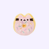 The Donut Mini Squisheen plush in front of a light purple background. The plush is a donut with cat ears, pink frosting and white, purple, teal and yellow sprinkles in the center. Among the sprinkles are four feet stubs facing forward, with a donut hole in the middle. By the ears, Pusheen’s head stripes, eyes and mouth are embroidered into the plush on top of the frosting, the head stripes a dark pink and the eyes and mouth in black.  There are black felt whiskers sticking out from the plush.