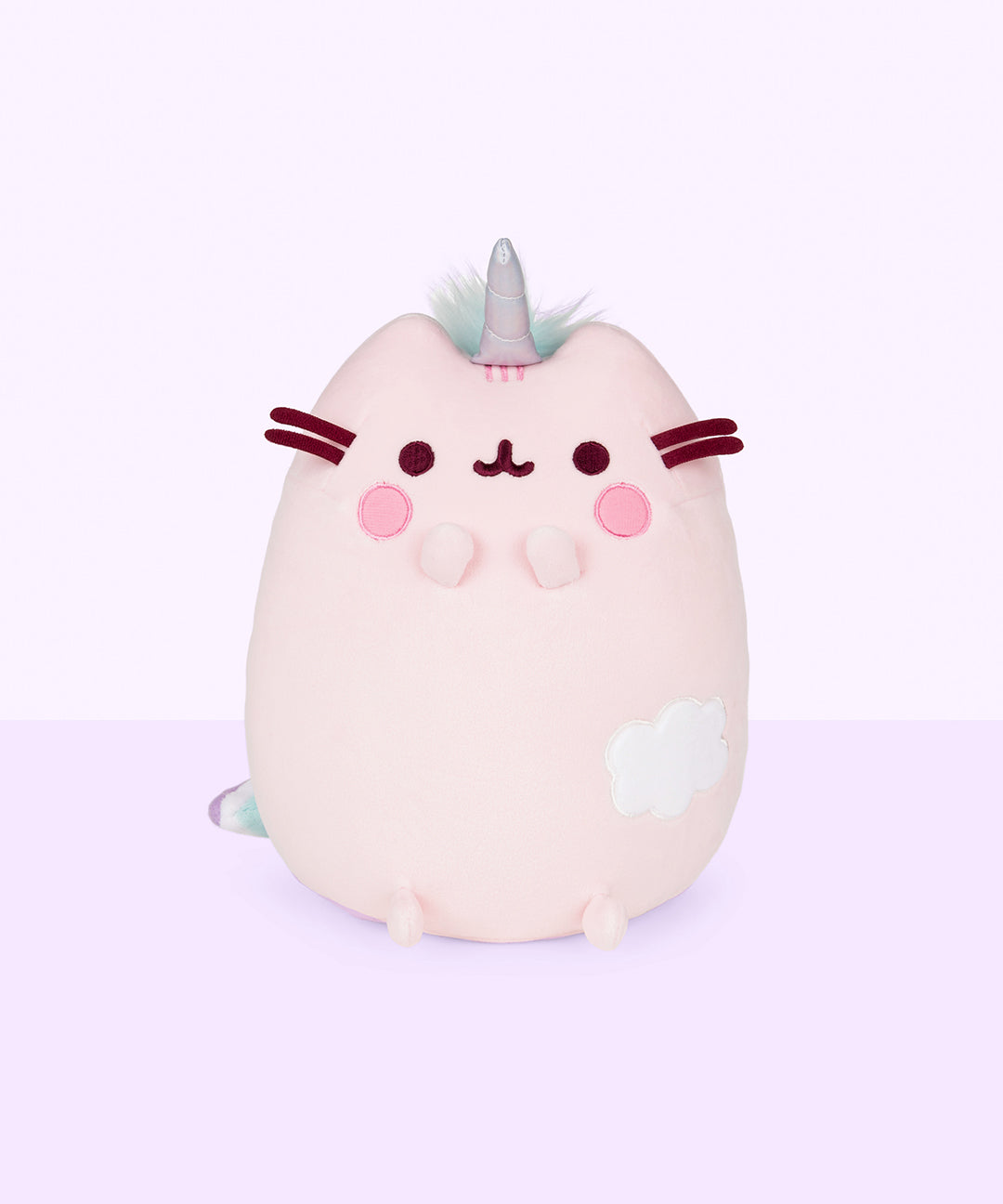 The Dreamy Pusheenicorn Squisheen in front of a light purple background. The plush features a light pink Pusheenicorn sitting upright with a unicorn horn in between her ears, circular blush marks, and an embroidered cloud on the lower right of her front body. Dreamy Pusheenicorn has two nub paws up in the air and two nub paws at the bottom. The embroidery on her eyes, mouth and felt whiskers are a dark magenta, while her head stripes are embroidered in pink.
