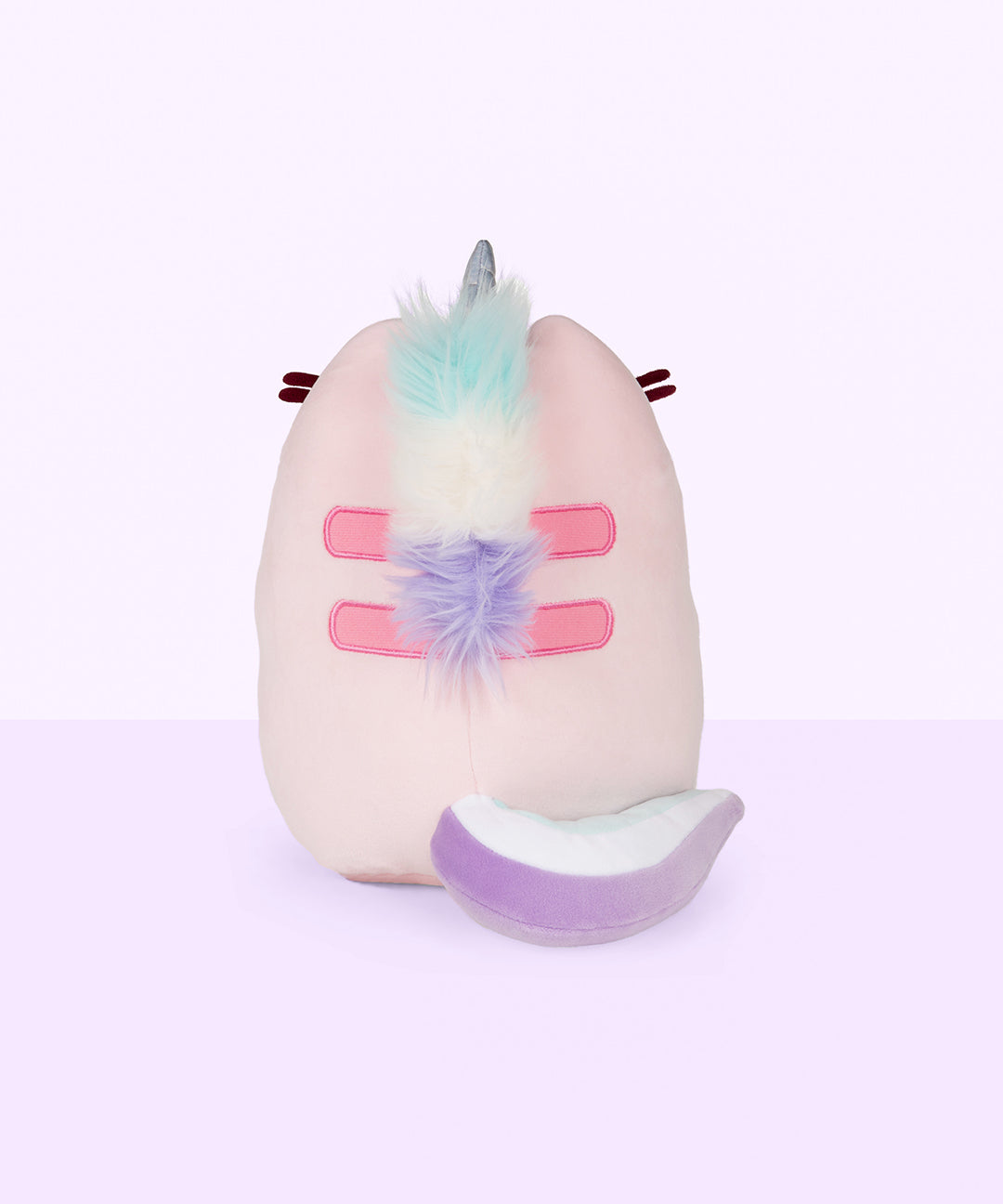 Back view of the Dreamy Pusheenicorn Squisheen in front of a light purple background. There is a multicolor mane directly behind Dreamy Pusheenicorn’s horn, colored mint, white and purple, going over Pusheen’s Back stripes and ending at the bottom stripe. Pusheen’s back stripes are embroidered in pink. Dreamy Pusheenicorn has a multicolor plush tail that matches her mane and curls around the bottom of her body to the right.