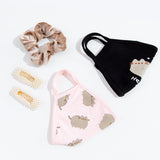 The Pusheen Hey Face Mask and the Pusheen Patterned face mask folded in half, laying besides each other and other accessories such as a scrunchie and two rectangular hair barrettes. 