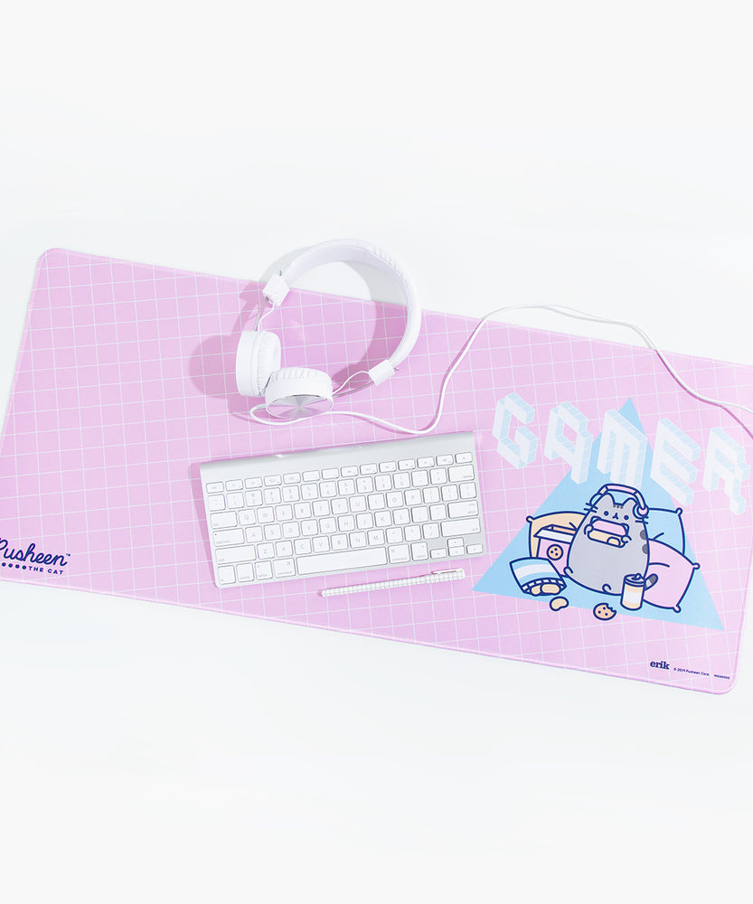Angled view of a pink rectangular desk mat with rounded edges, with white headphones, keyboard and a pen on top. The design on the mat features an angled white line grid pattern and a Gamer Pusheen pattern on the right hand side. Pusheen is wearing headphones and holding a controller while surrounded by pillows and snacks, in front of a blue triangle. The word 'GAMER' is above Pusheen in a white and light blue isometric pixel font .