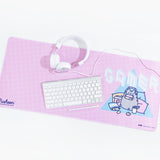 Angled view of a pink rectangular desk mat with rounded edges, with white headphones, keyboard and a pen on top. The design on the mat features an angled white line grid pattern and a Gamer Pusheen pattern on the right hand side. Pusheen is wearing headphones and holding a controller while surrounded by pillows and snacks, in front of a blue triangle. The word 'GAMER' is above Pusheen in a white and light blue isometric pixel font .