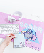Close up of the Pusheen Gaming Desk mat with items on top, including while headphones, a keyboard, and a hand writing a note note on a notepad. The Gaming Pusheen design on the desk mat is still visible.