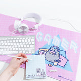 Close up of the Pusheen Gaming Desk mat with items on top, including while headphones, a keyboard, and a hand writing a note note on a notepad. The Gaming Pusheen design on the desk mat is still visible.