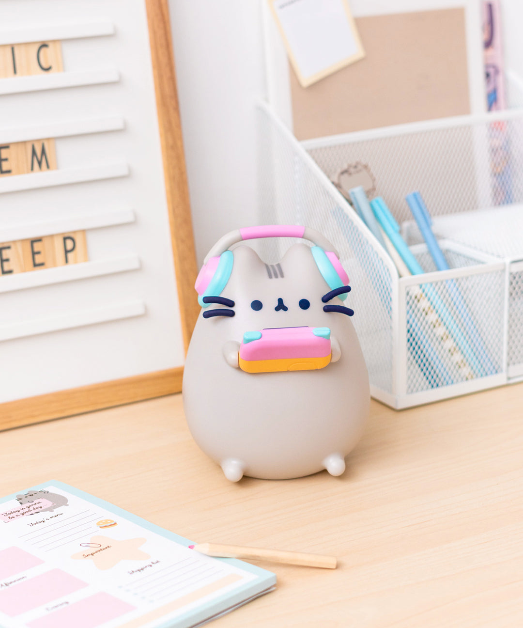 Front view of Gaming Pusheen Lamp. The grey tabby cat is holding a blue, pink, and orange gaming device in her paws while a pair of pink and blue headphones sit atop her ears. The cat’s facial features are navy blue while her head stripes are dark grey.  