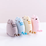 All four Mini Squisheen Plush lined up against one another, facing the left. There are two darker stripes embroidered on Pusheen’s side, and her striped tail sticks out from the side. The color lineup starts with grey, blue, yellow, and ends with pink.