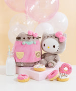 The Pusheen and Hello Kitty collaboration plush sit together, A yellow background and pink and white balloons behind them, and donuts in front of them. A milk jug besides Pusheen features two striped straws.