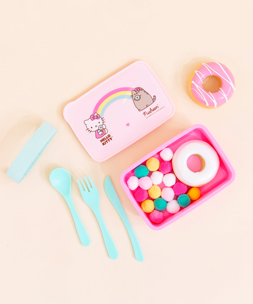 Top view of the a pink lunchbox with it’s lid placed above it, a teal knife, fork, spoon and rubber band placed neatly to the left. The lunch box lid features Hello Kitty holding a donut on the left with the text ‘Hello Kitty’ underneath her, Pusheen wearing a hair bow and holding a donut on the right with the text ‘Pusheen’ underneath, and a rainbow in the middle connecting the two with a small pink heart underneath it. Multicolor pom poms and a white glazed donut are inside the box.