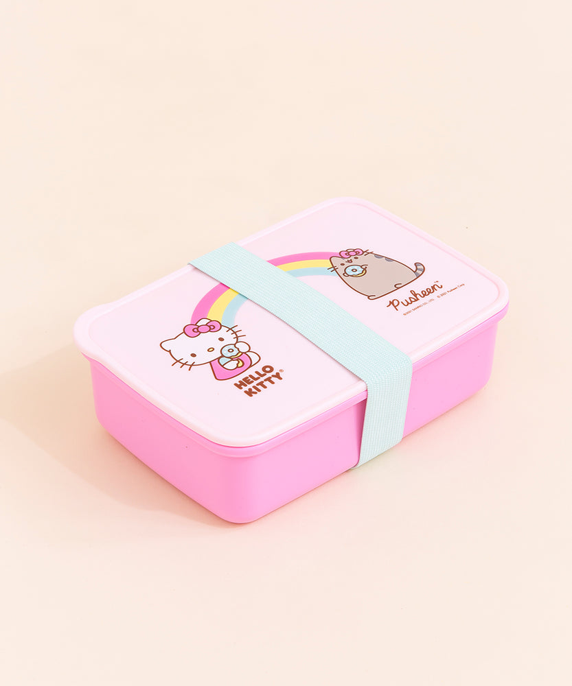 The Hello Kitty x Pusheen Lunchbox closed, with the teal band keeping the lunch box closed. The lid is a lighter pink then the box, and the lunch box is a rounded rectangle. The lunchbox is placed in front of a cream background.