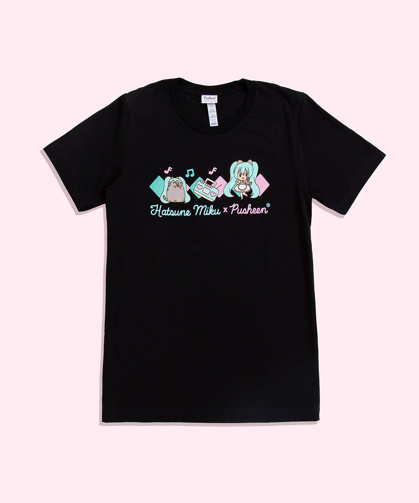 A short sleeve black shirt featuring a graphic in the middle in front of a light pink background. The graphic features Pusheen wearing a Miku wig, a boom box, and Miku dressed in a pink poofy dress with cat ears. The both of them holding a microphone and singing on top of a pink and teal diamond pattern, the Hatsune Miku x Pusheen collaboration logo underneath them.