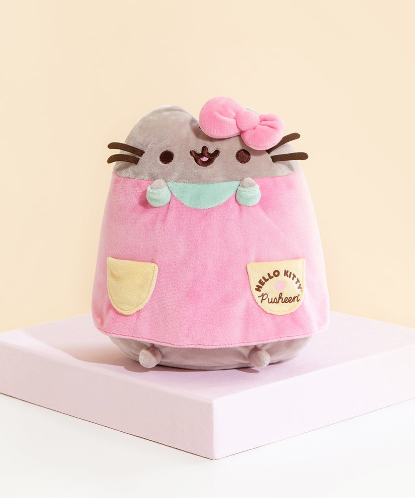 A sitting Pusheen Plush wearing a pink dress with yellow pockets, mint sleeves and a collar, as well as Hello Kitty's bow on top of her head. The Hello Kitty x Pusheen collaboration logo is present on the right dress pocket.