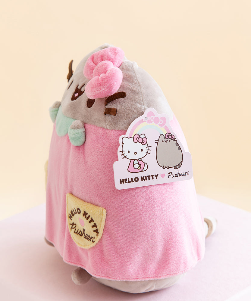 Side view of the plush, showing the product tag for the item. The tag features artwork of Hello Kitty sitting across from Pusheen, who is wearing Hello Kitty's bow. A rainbow with Hello Kitty's bow connects the two.
