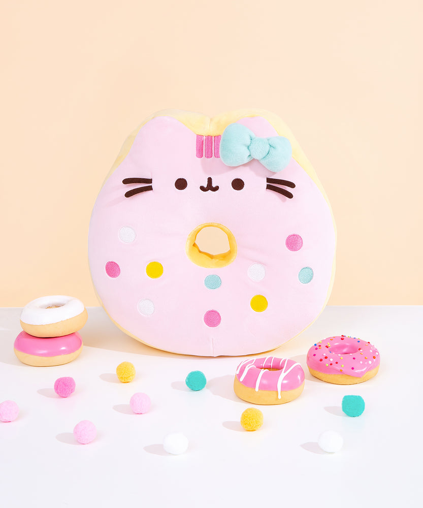 Front view of Pusheen side of Hello Kitty x Pusheen Donut Plush on a white surface in front of a yellow background. Pusheen the Cat takes the form of a pink glazed donut accented with multicolor blue, white, yellow, and pink sprinkles and a light blue bow to match Hello Kitty’s pink bow on the reverse side. 