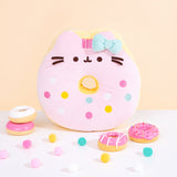 Front view of Pusheen side of Hello Kitty x Pusheen Donut Plush on a white surface in front of a yellow background. Pusheen the Cat takes the form of a pink glazed donut accented with multicolor blue, white, yellow, and pink sprinkles and a light blue bow to match Hello Kitty’s pink bow on the reverse side. 