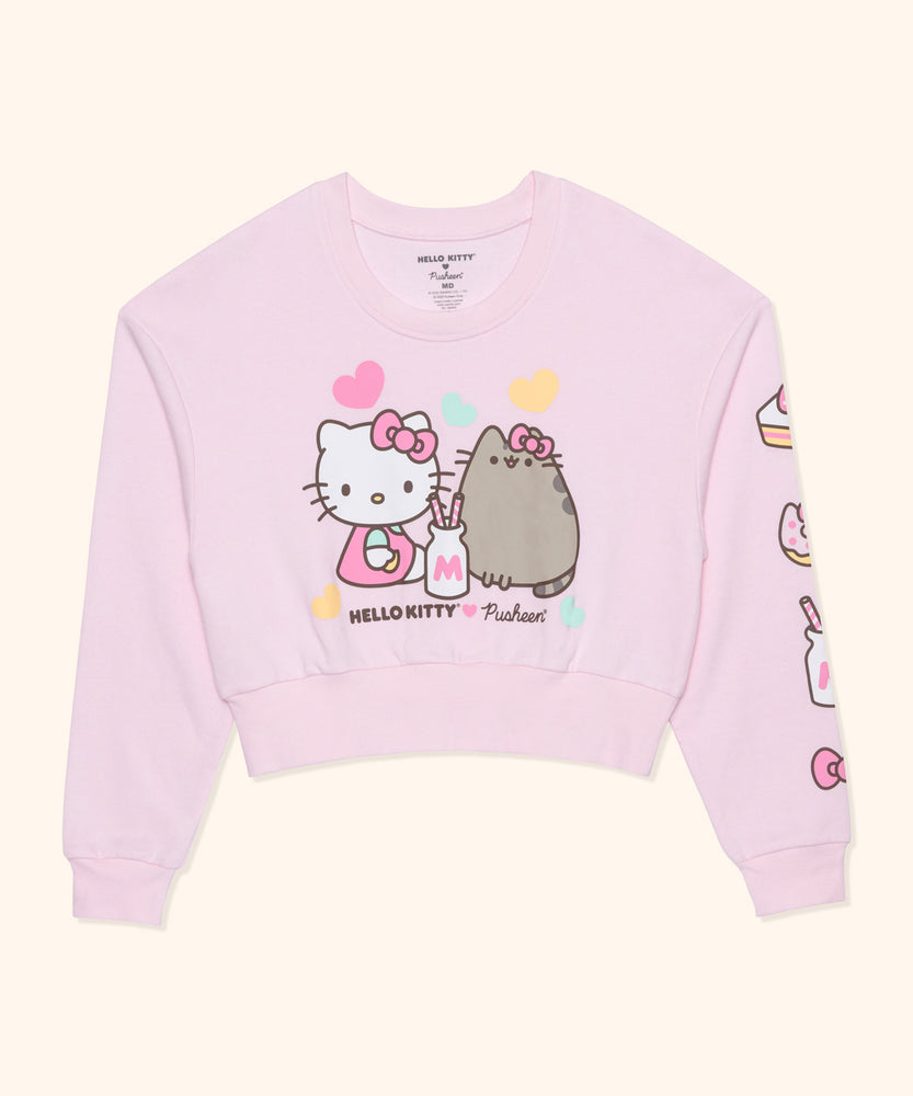 A long sleeve light pink sweatshirt laid out flat on top a light yellow surface. In the center of the cropped ladies sweatshirt is a graphic of Hello Kitty and Pusheen sharing a milkshake while surrounded by multi-color hearts. Under the center graphic is the Hello Kitty x Pusheen collaboration logo.Down the left sleeve of the sweatshirt are graphics of a slice of cake, donut with cat ears, a milk jar with two straws, and Hello Kitty's signature bow.