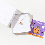 The gold charm necklace in a square white jewelry box. A square card insert is next to the box, featuring artwork of the charm.