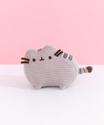 Front view of plush. Knit Pusheen Plush sits atop a white surface in front of a light pink background. The mini plush has a small grey crocheted look to it. 
