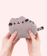 Model holds mini knit plush to demonstrate how small the plush is. Next to model’s right hand, the crocheted grey and brown tail extends off the body of the plush. 
