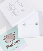 Front view of Pusheen Lazy Stud Earrings, which are unmatched loaf poses of Pusheen the Cat. The earrings on the left are sterling silver and on the right are the rose gold vermeil earrings. Behind the earrings sits the white packaging case that the earrings come in. 