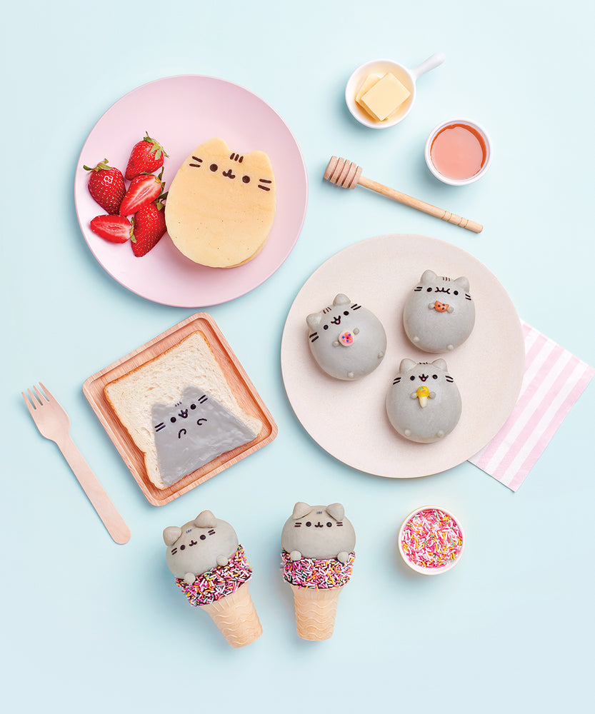 A photo of various Pusheen treats from the cookbook, arranged on plates on top of a light blue surface. Some of the baked goods include Pusheen Pancakes, Pusheen on toast, a dome cookie of Pusheen holding various mini food, and Pusheen peeking out of a sprinkle cake cone.