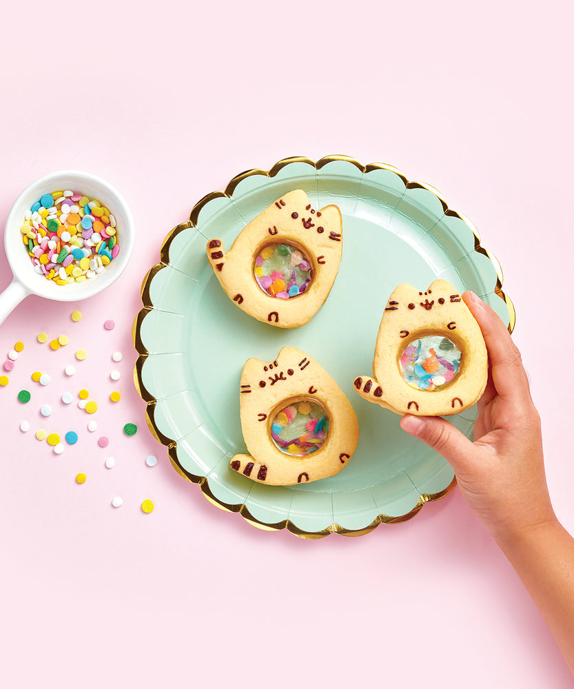 Top view of a trio of Pusheen cookies with a see-through stomach with sprinkles inside. A model’s hand holds one of the cookies above the mint and gold plate, and a scoop of sprinkles has slightly spilled to the left.
