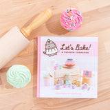 The Pusheen Cookbook laying flat on top of a wooden surface, surrounded by a wooden rolling pin and a pink and mint frosted cupcake. The spine of the book is pink, and by the title of the book is an illustration of Pusheen in a white apron and a chef’s hat.