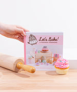 A hand holdind the corner of the Pusheen cook book, standing on top of a wooden surface surrounded by a rolling pin and a pink frosted cupcake.
