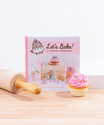 A square hardcover book standing on top of a wooden board, surrounded by a rolling pin, a pink cupcake, and a white pedestal. The book reads ‘Let’s Bake! A Pusheen Cookbook’ and features a photo displaying all the Pusheen treats you can bake with the book. The author names Claire Belton and Susanne Ng are noted in pink at the bottom.