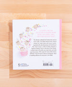 View of the back cover of the cook book on top of a wooden surface. The back cover features Pusheen-shaped marshmallows erupting from a pink cupcake cup alongside regular marshmallows and multicolor stars. The back reads ‘A Color and fun collection of PUsheen-themed recipes for cookies, cakes and other delicious treats’ in pink, with more back copy in dark brown. The publisher’s information is noted on the lower left, and the barcode is noted on the lower right.