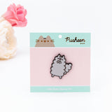 The Stormy pin in it’s packaging, laying on a white floor, a pair of pink and white peonies partially visible in the top left corner. The packaging is a square cardboard backing, a pink square in the middle and mint banners on the top and bottom. The top banner features the Pusheen Shop logo, and the bottom banner says ‘Little Sister Stormy Pin’ in white.